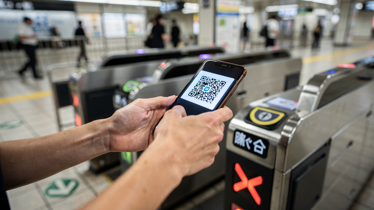A person's hands holding a smartphone, blurred in motion, while reaching out to scan a QR code displayed on a digital screen at a Tokyo metro station ticket gate, capturing a moment of digital interaction in a bustling urban environment.