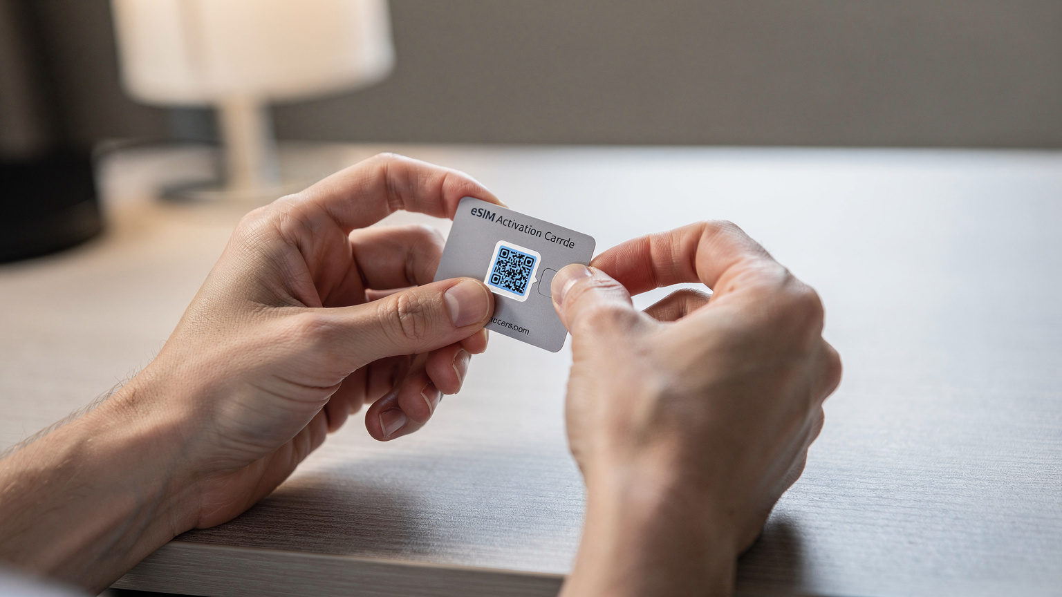 A close-up view of a person's hands carefully peeling a QR code sticker from a small eSIM package, preparing for activation in a well-lit hotel room.