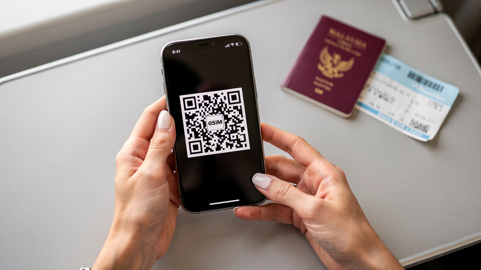Tangan seorang wanita sedang memegang telefon pintar yang menunjukkan kod QR eSIM di skrin, bersedia untuk diimbas, dengan latar belakang pasport Malaysia dan tiket penerbangan.