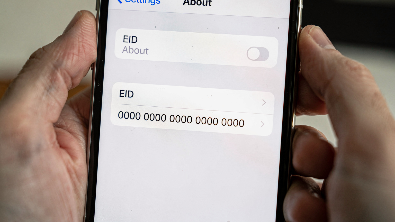 A close-up shot of an iPhone screen displaying the 'About' section in Settings, with the 'EID' field highlighted, indicating eSIM compatibility. The hands holding the phone are gently blurred in the foreground.