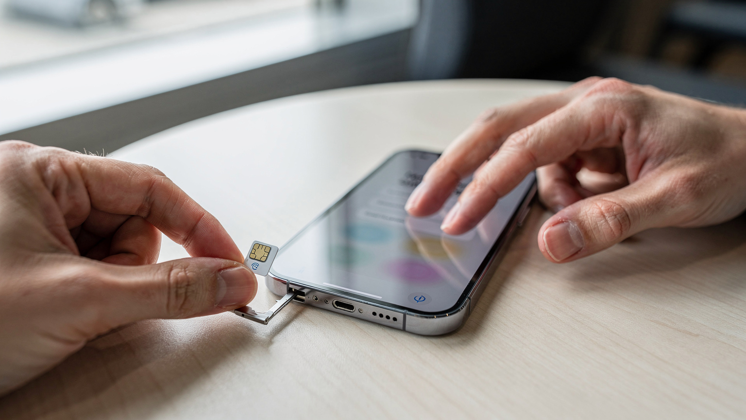 A close-up of a traveler's hands carefully aligning a physical SIM card next to an iPhone with an eSIM profile visible on the screen, illustrating the contrast between old and new connectivity methods. The setting is a clean, modern airport lounge table.