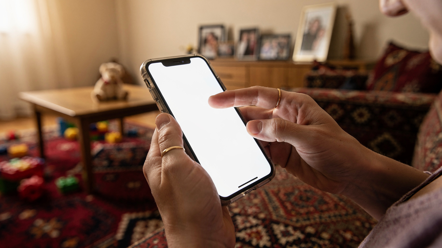 Bir annenin eli, eski iPhone'unun ekranında 'Tüm İçerikleri ve Ayarları Sil' seçeneğine dokunmak üzere. Arka planda, çocukların oyuncakları ve aile fotoğrafları belirsiz bir şekilde görünüyor, evin sıcak ortamını yansıtıyor.