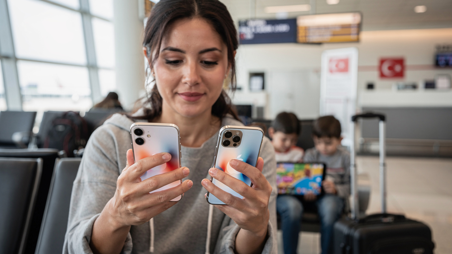 Bir annenin elinde iki iPhone, biri eski diğeri yeni model, iOS Hızlı Aktarım ekranında adım adım eSIM aktarımını gerçekleştiriyor. Arkada, uçak kalkışına hazırlanan bir aile, çocuklarının tabletlerine oyunlar yüklüyor.