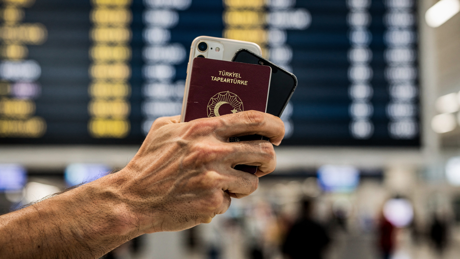 Bir seyahat blog yazarının pasaportunu ve iPhone'unu tuttuğu bir el, arkada bir havalimanı kalkış panosu.