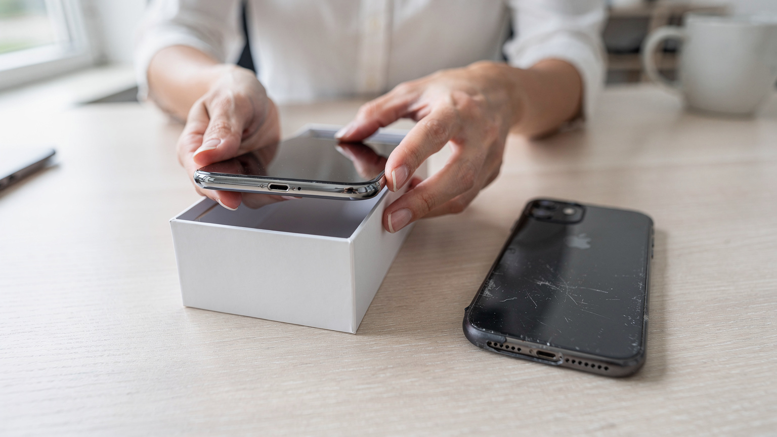 Nahaufnahme der Hände einer Frau, die vorsichtig ein glänzendes neues iPhone aus seiner Verpackung hebt, während daneben ein altes iPhone liegt, bereit für die Datenübertragung.