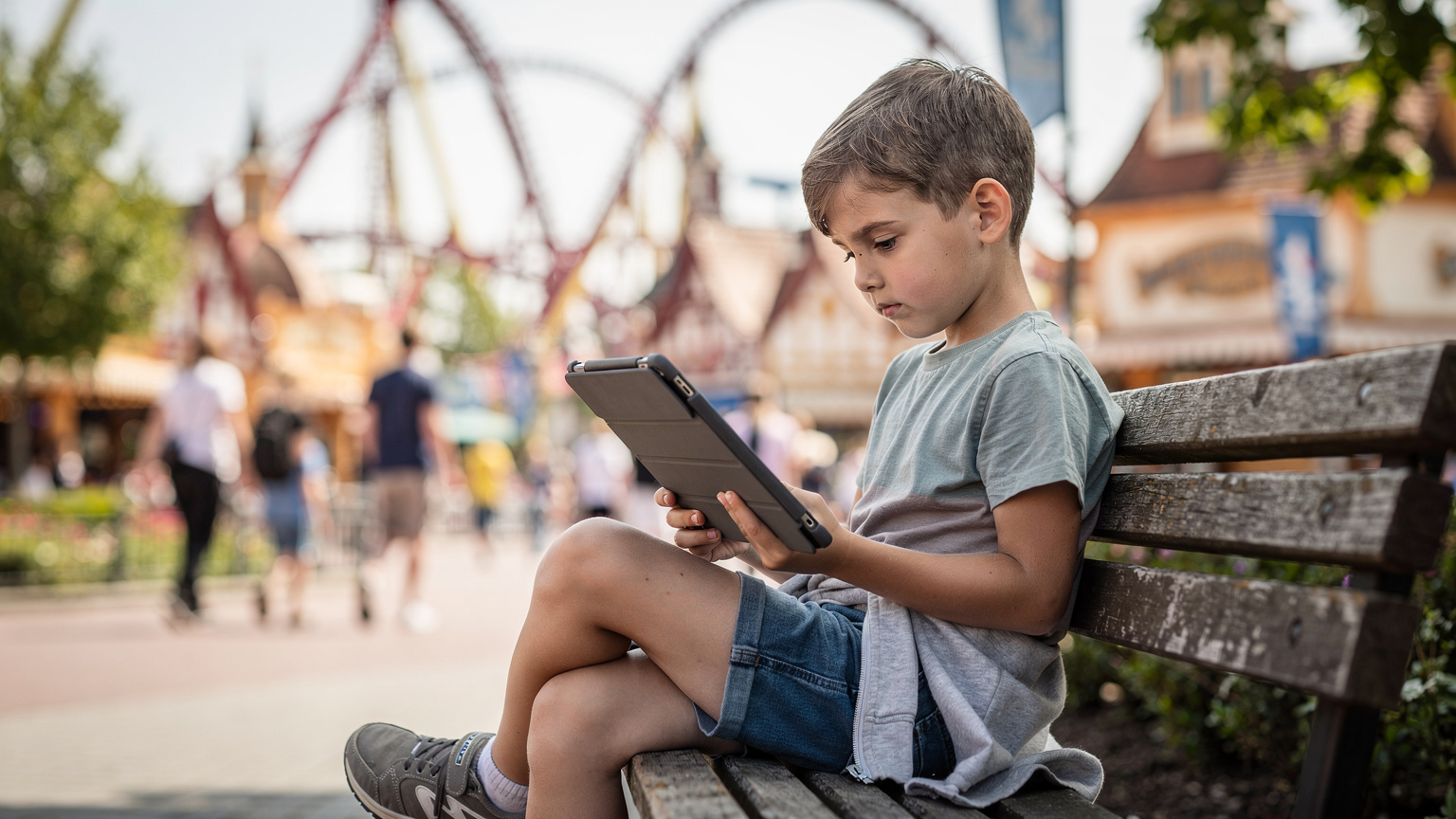 Ein 10-jähriges Kind sitzt auf einer Bank in einem Themenpark in Rust, vertieft in sein iPad, das über eine eSIM mit dem Internet verbunden ist, während es eine Zeichentrickserie schaut.
