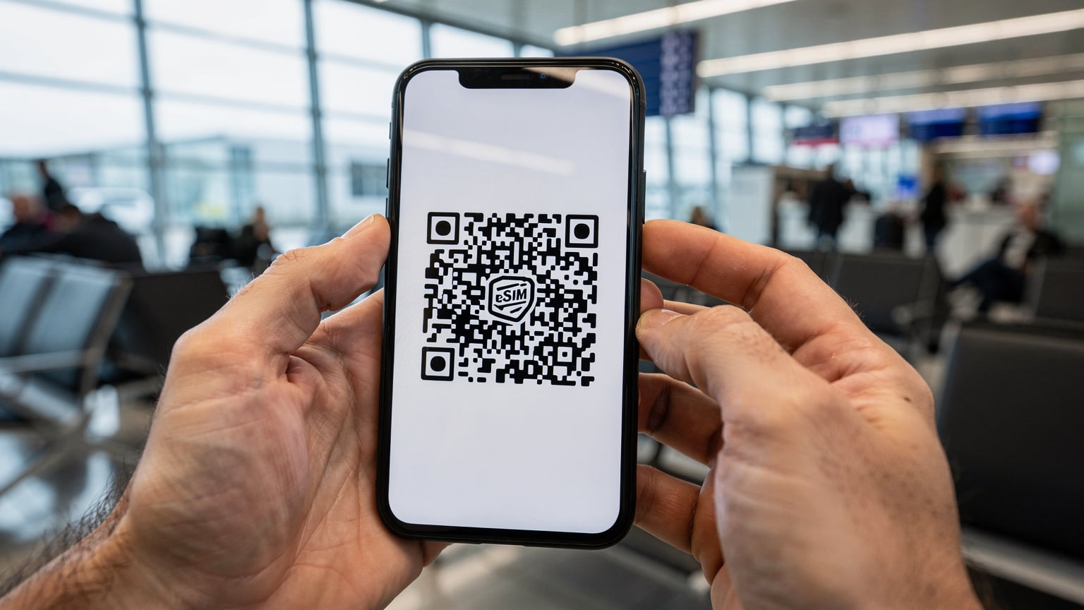Eine Nahaufnahme eines Smartphones, das einen QR-Code für die eSIM-Aktivierung anzeigt, mit unscharfen Händen, die den Bildschirm halten. Im Hintergrund ist ein geschäftiger Flughafen-Warteraum in München zu sehen.