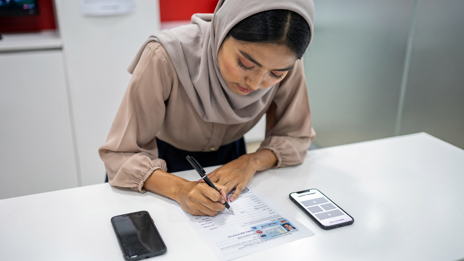 Seorang wanita muda Indonesia sedang mengisi formulir di meja layanan pelanggan GraPARI Telkomsel, dengan ID card dan ponsel di sampingnya, menunjukkan proses registrasi yang teliti.