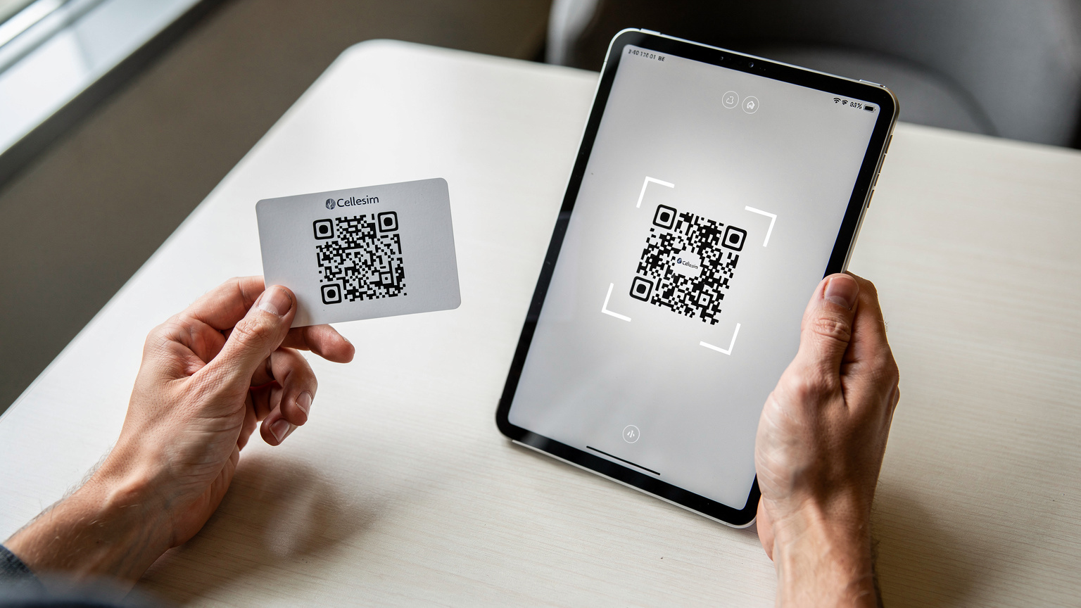 Nahaufnahme von Händen, die ein iPad halten und den Bildschirm berühren. Eine QR-Code-Scanning-Ansicht ist geöffnet, während eine andere Hand eine ausgedruckte Cellesim-eSIM-Karte mit einem QR-Code hält, der gescannt werden soll.