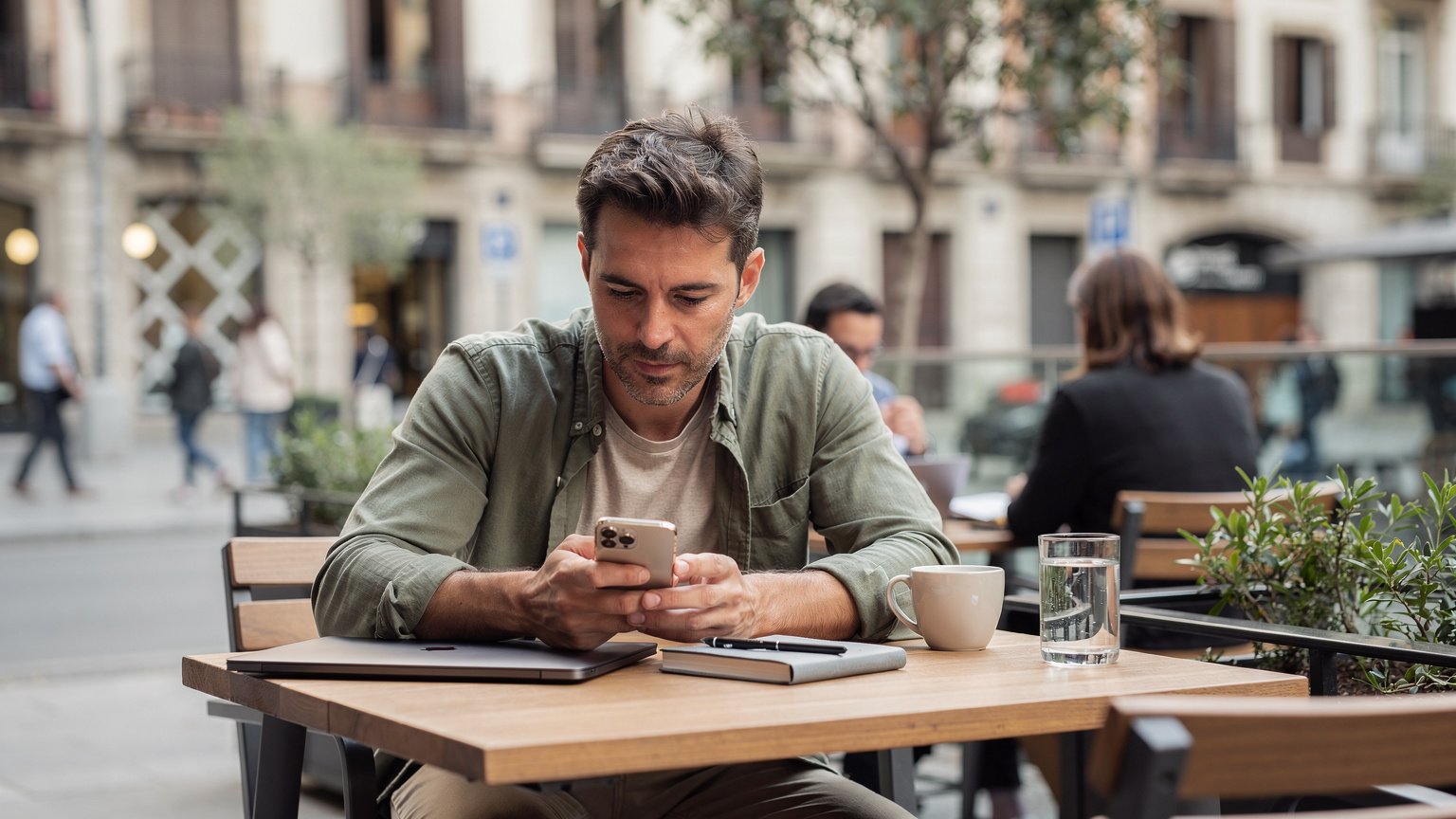 Un nómada digital trabajando en su iPhone en una mesa al aire libre en un co-working en Barcelona, con gente pasando al fondo. La atmósfera es relajada y productiva, bajo una luz natural brillante.