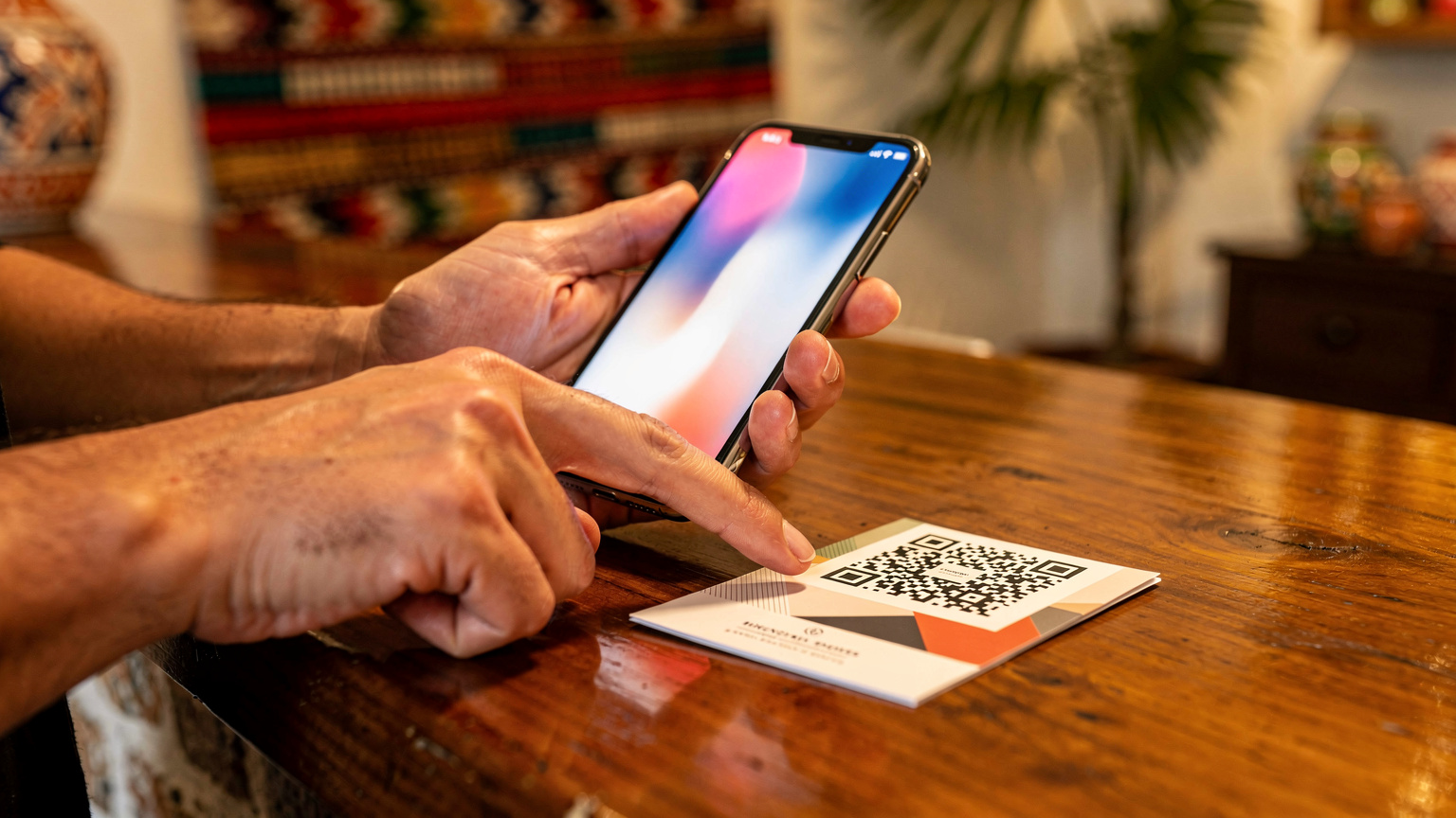 Un primer plano de manos humanas, una sosteniendo un iPhone y la otra apuntando a un código QR en un folleto, en el mostrador de un hotel boutique en Cartagena, Colombia. La luz es cálida y acogedora.