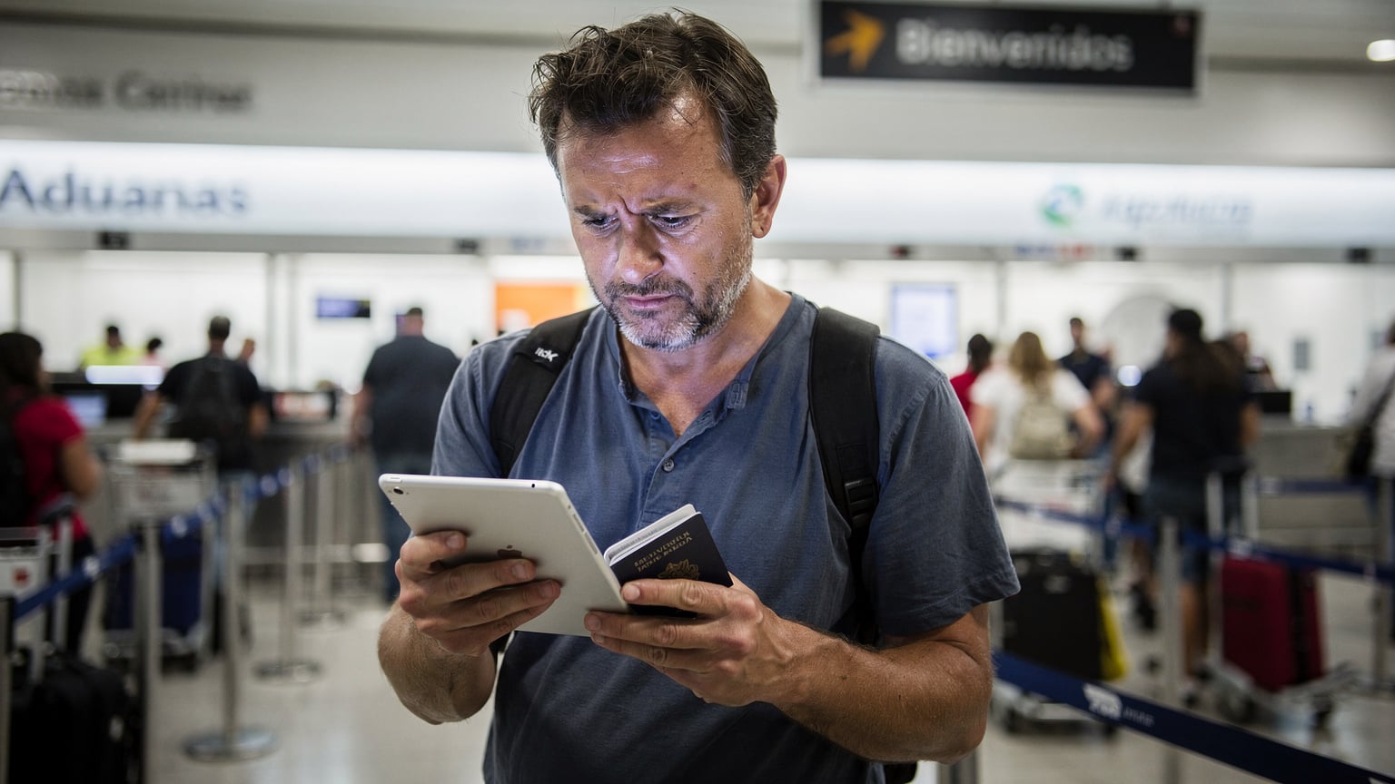 Un viajero frustrado en la aduana de un aeropuerto, sosteniendo su iPad y un pasaporte, con una expresión de preocupación mientras mira la pantalla que muestra un error de conexión, en una escena de aeropuerto genérico pero con detalles culturales latinos, como un cartel en español.