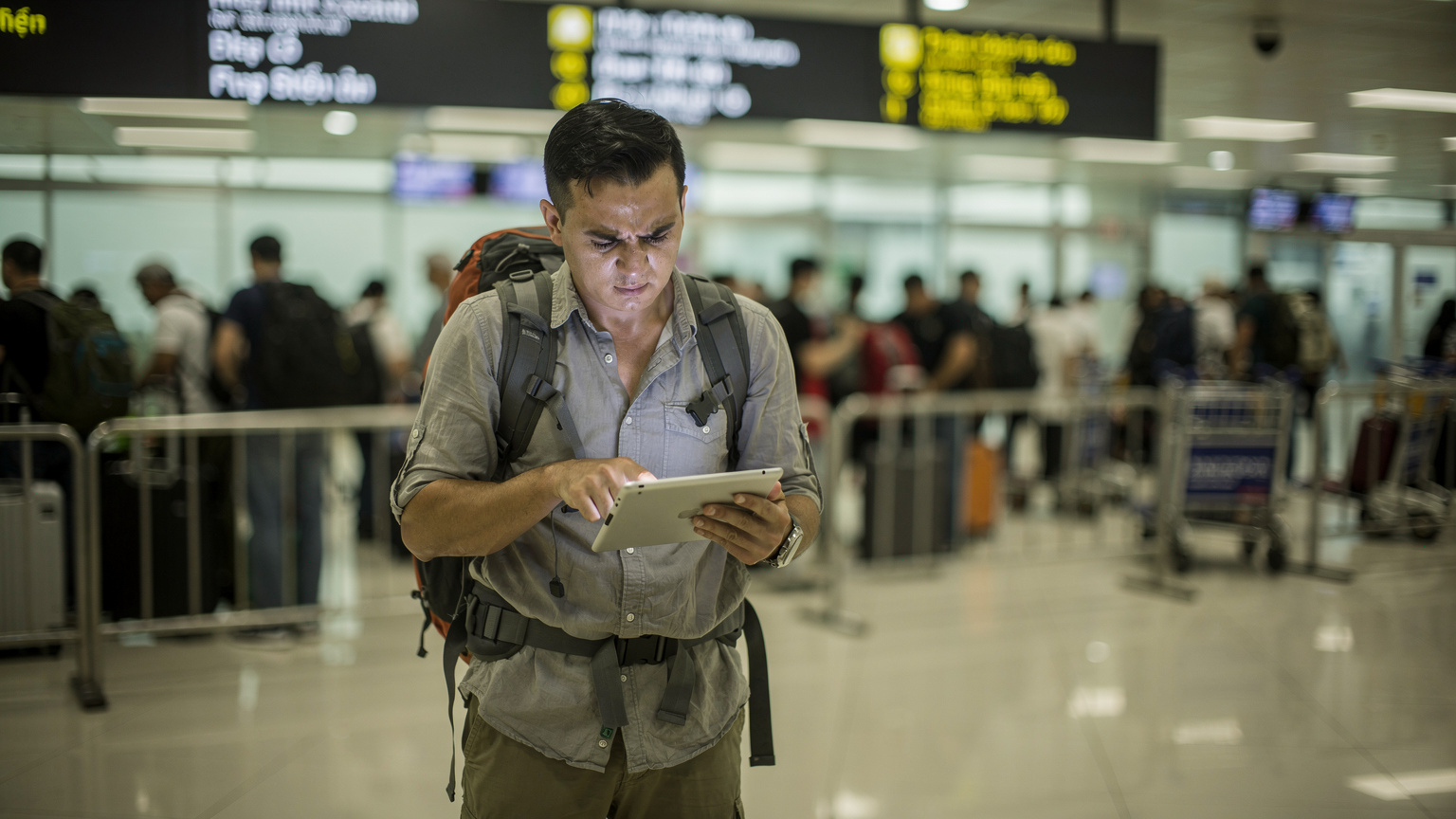 Un viaggiatore sta aspettando in fila all'aeroporto di Hanoi, con un'espressione di frustrazione mentre cerca di connettersi a una rete Wi-Fi instabile sul suo iPad, con lo sfondo sfocato di un'area di arrivi affollata.
