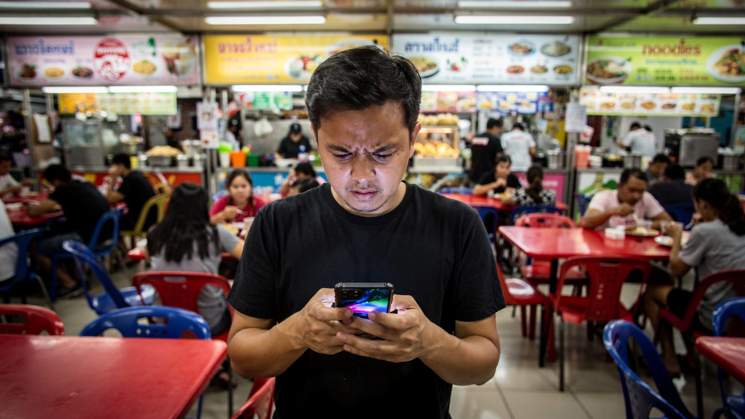 O scenă aglomerată dintr-un food court în Bangkok, unde un călător pare stresat, uitându-se la telefonul său deschis, cu o expresie îngrijorată, în timp ce alți oameni mănâncă în jurul său.