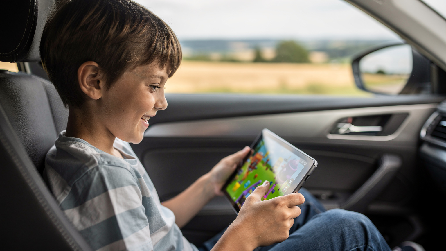 Un enfant souriant, absorbé par un jeu sur sa tablette dans le siège arrière d'une voiture, illustrant l'importance de la connectivité mobile pour divertir les jeunes voyageurs, avec un paysage flou de campagne défilant par la fenêtre.