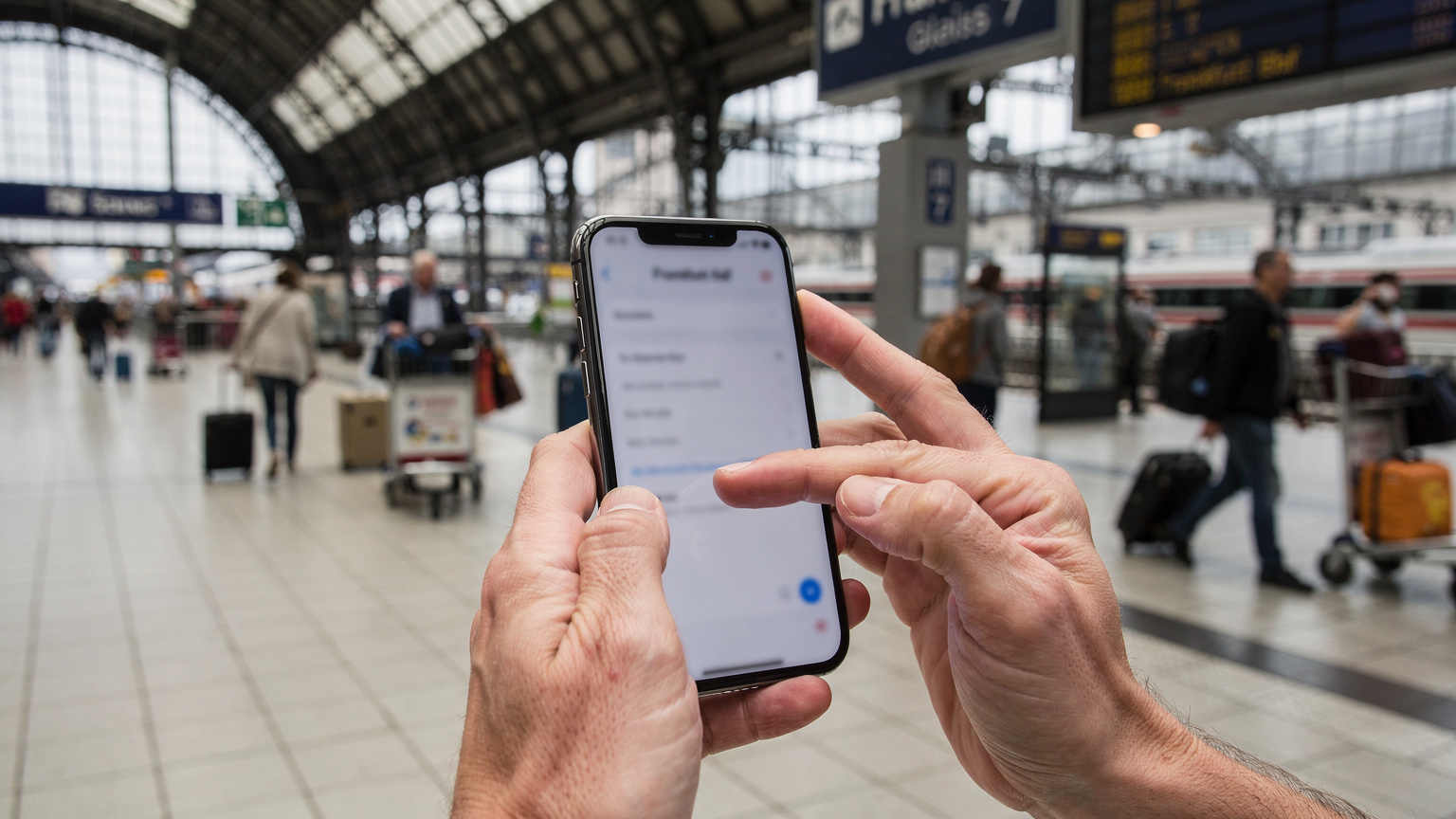 Mână de părinte ajustând setările de date pe un smartphone în timpul unei călătorii, cu un fundal estompat de gară aglomerată din Frankfurt, Germania.