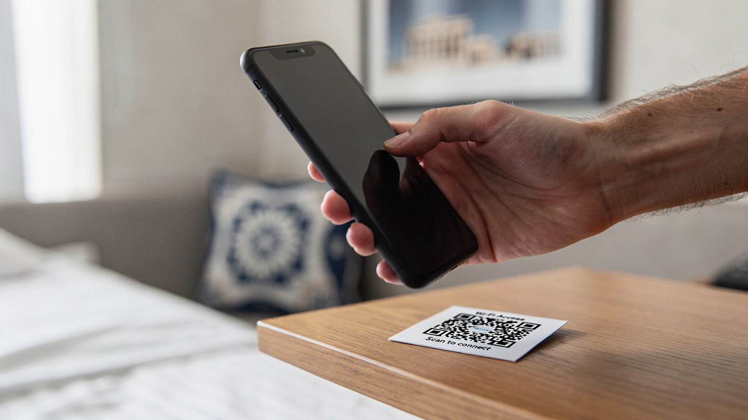 Mână masculină ținând un smartphone, pe ecranul căruia este vizibilă o interfață generică de setări pentru rețea, în timp ce scanează un cod QR de pe o fișă, într-un interior luminos de cameră de hotel.