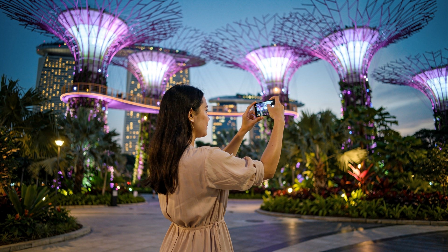 Seorang wanita muda Indonesia sedang mengambil foto suaminya dengan latar belakang Supertree Grove di Gardens by the Bay, Singapura, saat langit senja dengan lampu-lampu yang mulai menyala.