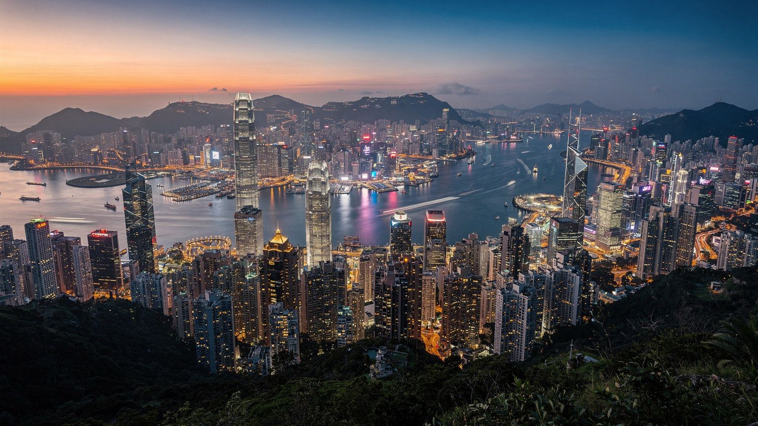 Pandangan udara dari Victoria Peak di Hong Kong, menampilkan cakrawala kota yang ikonik di bawah langit senja, dengan kapal-kapal kecil di Pelabuhan Victoria.