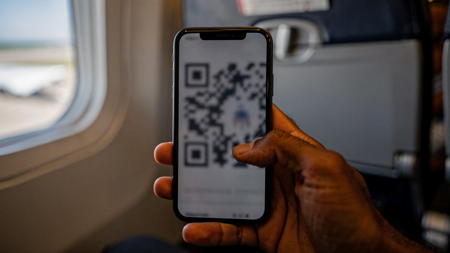 Tangan seorang pria Indonesia memegang ponsel dengan layar kode QR eSIM yang siap dipindai, di latar belakang kabin pesawat yang nyaman sebelum lepas landas.