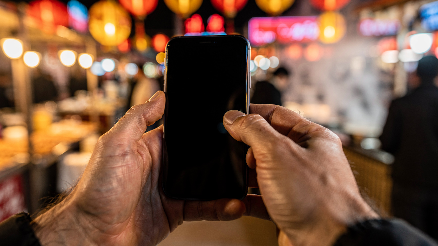 Primer plano de manos sosteniendo un smartphone, con el pulgar moviéndose por la pantalla, en un bullicioso mercado nocturno de Kashgar, China, con luces de neón y gente difusa de fondo.