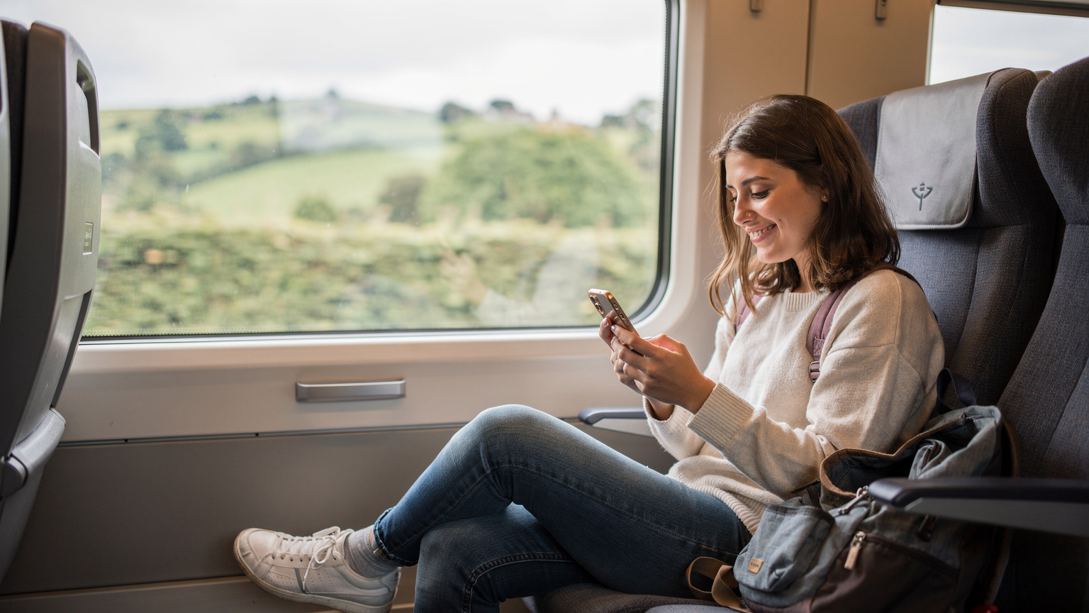 A young female student seated on a train in the UK, looking at her phone and smiling, with a backpack by her feet, indicating travel and connectivity.
