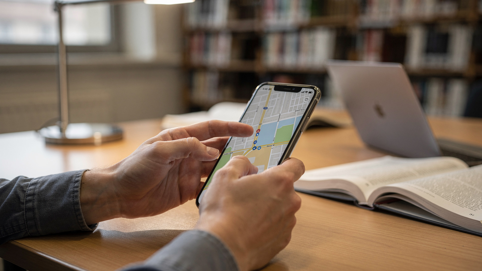 Close-up of a student's hands navigating a map application on a smartphone, with blurred academic books and a laptop in the background in a university library.