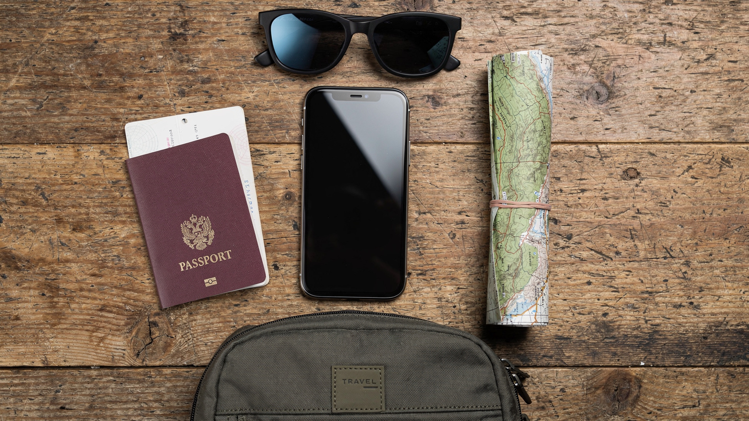 En oversiktlig flat-lay av reiseutstyr: en smarttelefon med eSIM-aktiveringsskjermen, et pass, en liten ryggsekk, et par solbriller og et fjellkart, alt på en rustikk treflate.