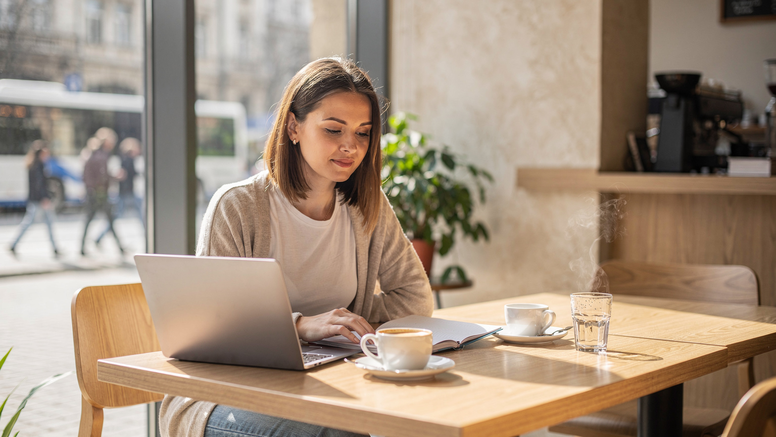 Un digital nomad stă într-o cafenea modernă și luminoasă din Chișinău, cu laptopul deschis și o cafea aburindă pe masă, concentrat pe ecran, în timp ce în fundal se observă activitatea urbană prin fereastră.
