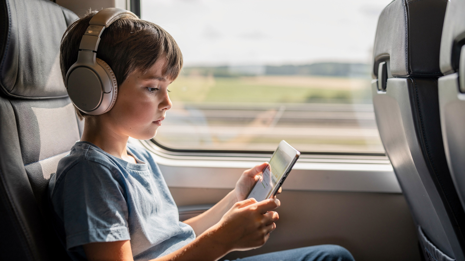 A child, aged about 9, with headphones on, looking intently at an iPad screen while sitting comfortably on a high-speed train, with blurred scenery passing outside the window.
