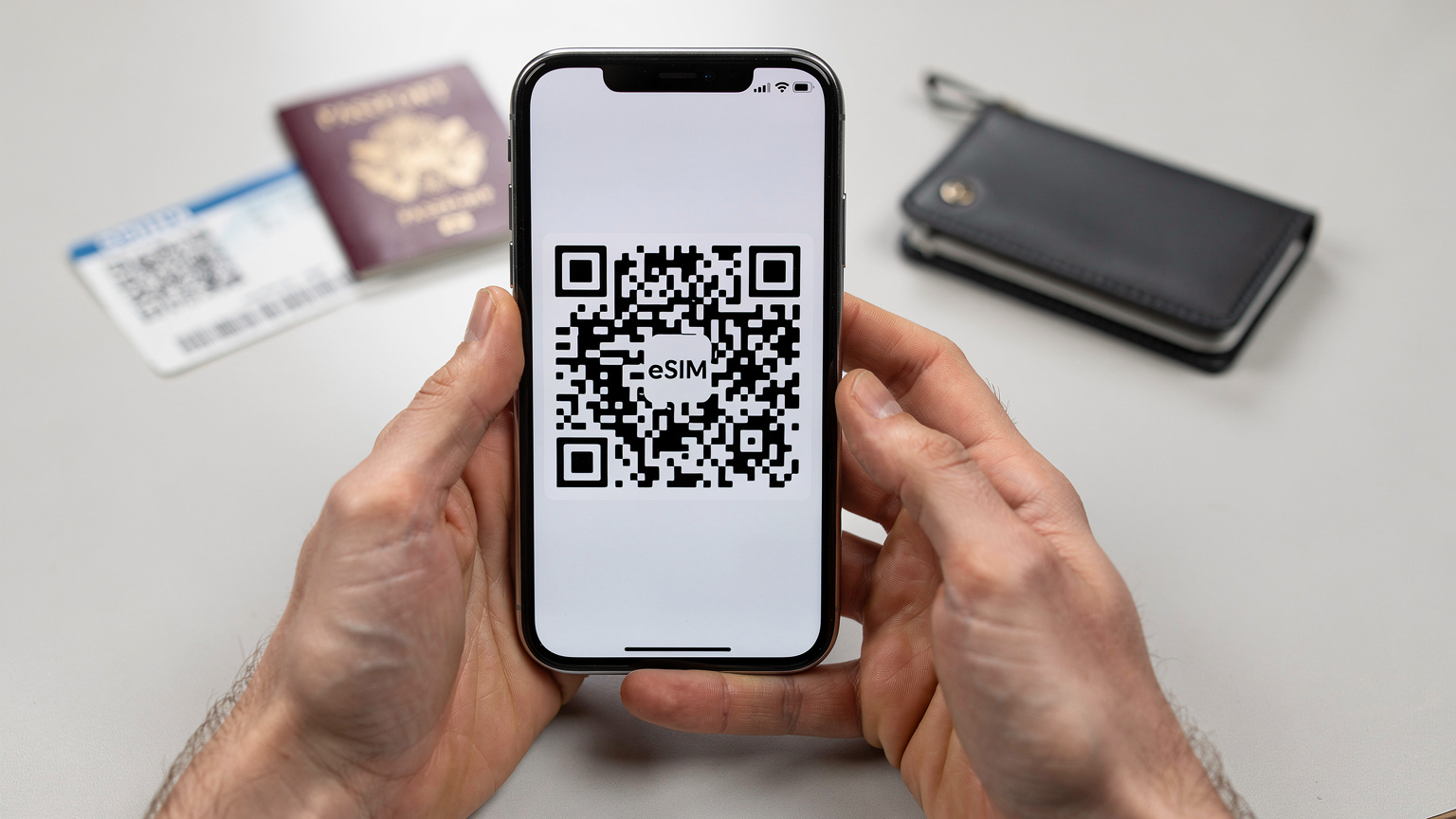 Close-up of human hands holding a smartphone, displaying a QR code on the screen, with a blurred passport and boarding pass in the background, implying travel preparation.