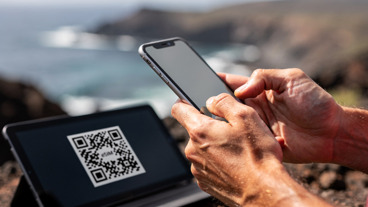 Close-up of human hands holding a smartphone, with a QR code displayed on a separate tablet screen for eSIM installation. The background is a blurred, natural Galápagos scene.