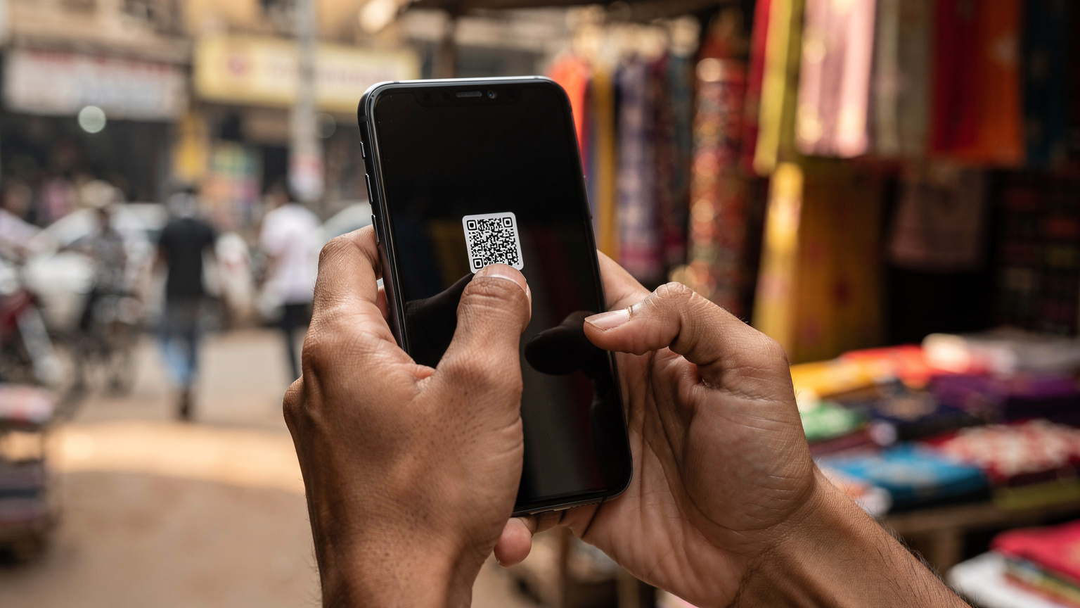 एक भारतीय यात्री के हाथों का क्लोज-अप, जो अपने स्मार्टफोन पर eSIM QR कोड स्कैन कर रहा है। पृष्ठभूमि में, एक हलचल भरे भारतीय बाजार की धुंधली झलक दिख रही है।