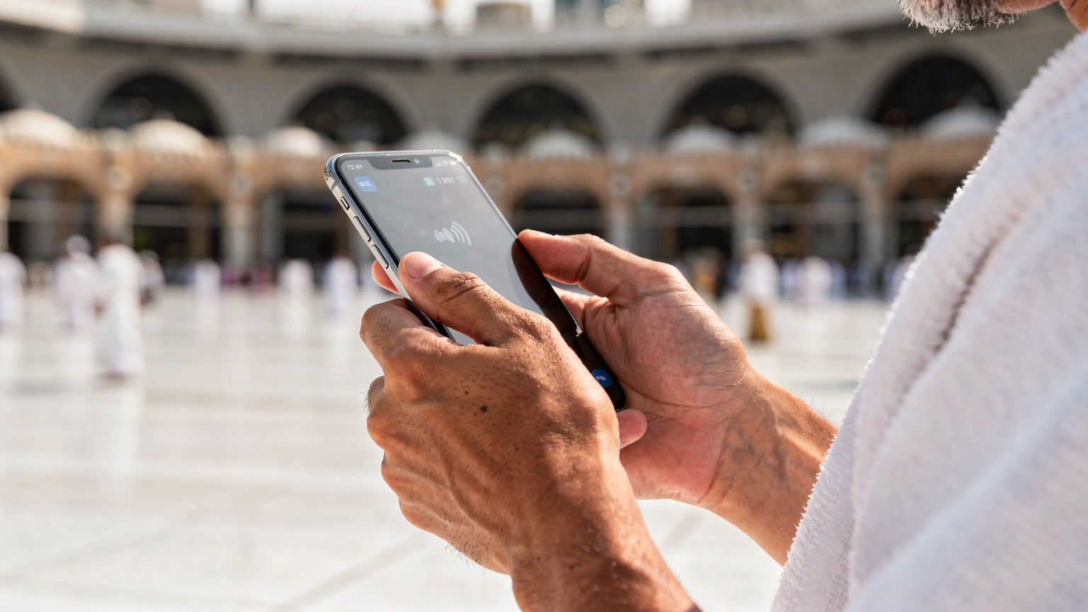Seorang jemaah lelaki Malaysia sedang memegang telefon pintar dengan skrin menunjukkan ikon rangkaian selular, di perkarangan Masjidil Haram di Makkah.