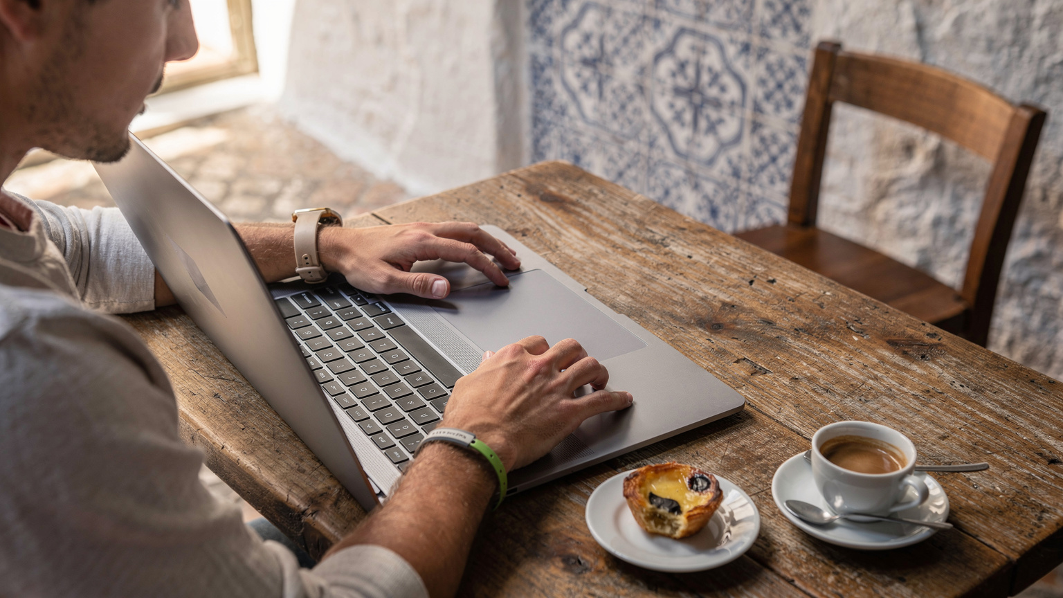 Uma pessoa digitando em um laptop em um café charmoso em Lagos, Algarve, com uma xícara de café e um pastel de nata na mesa, aproveitando a conexão Wi-Fi ou eSIM.