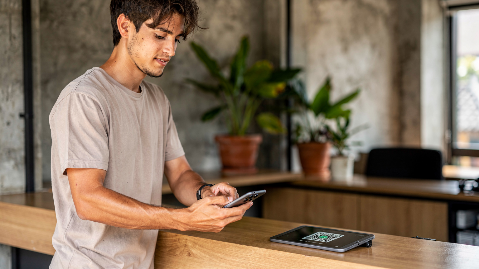 Seorang traveler dengan rambut sebahu, mengenakan kaus polos, sedang mencoba memindai kode QR eSIM di layar Samsung Galaxy S24 miliknya. Dia berdiri di depan meja resepsionis sebuah hostel dengan gaya industrial di kawasan Legian, Bali. Suasana pagi yang tenang dengan beberapa tanaman hias di latar belakang.