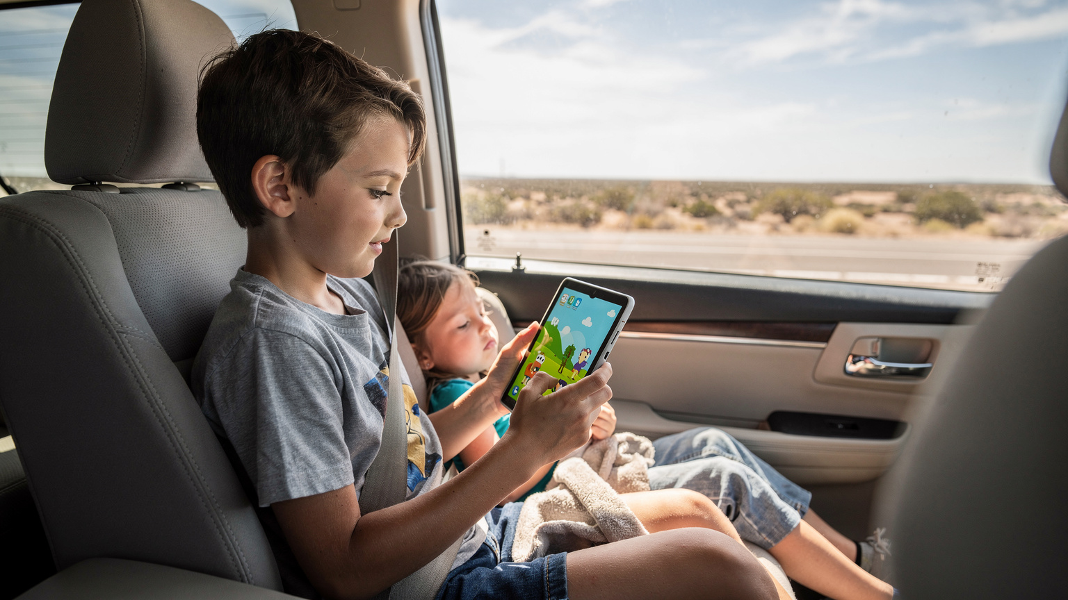 Un bambino di circa 10 anni è seduto sul sedile posteriore di un'auto a noleggio in Texas, con un tablet in mano che mostra un cartone animato di Disney+, mentre fuori dal finestrino scorre un paesaggio desertico. Un fratello più piccolo dorme accanto a lui.