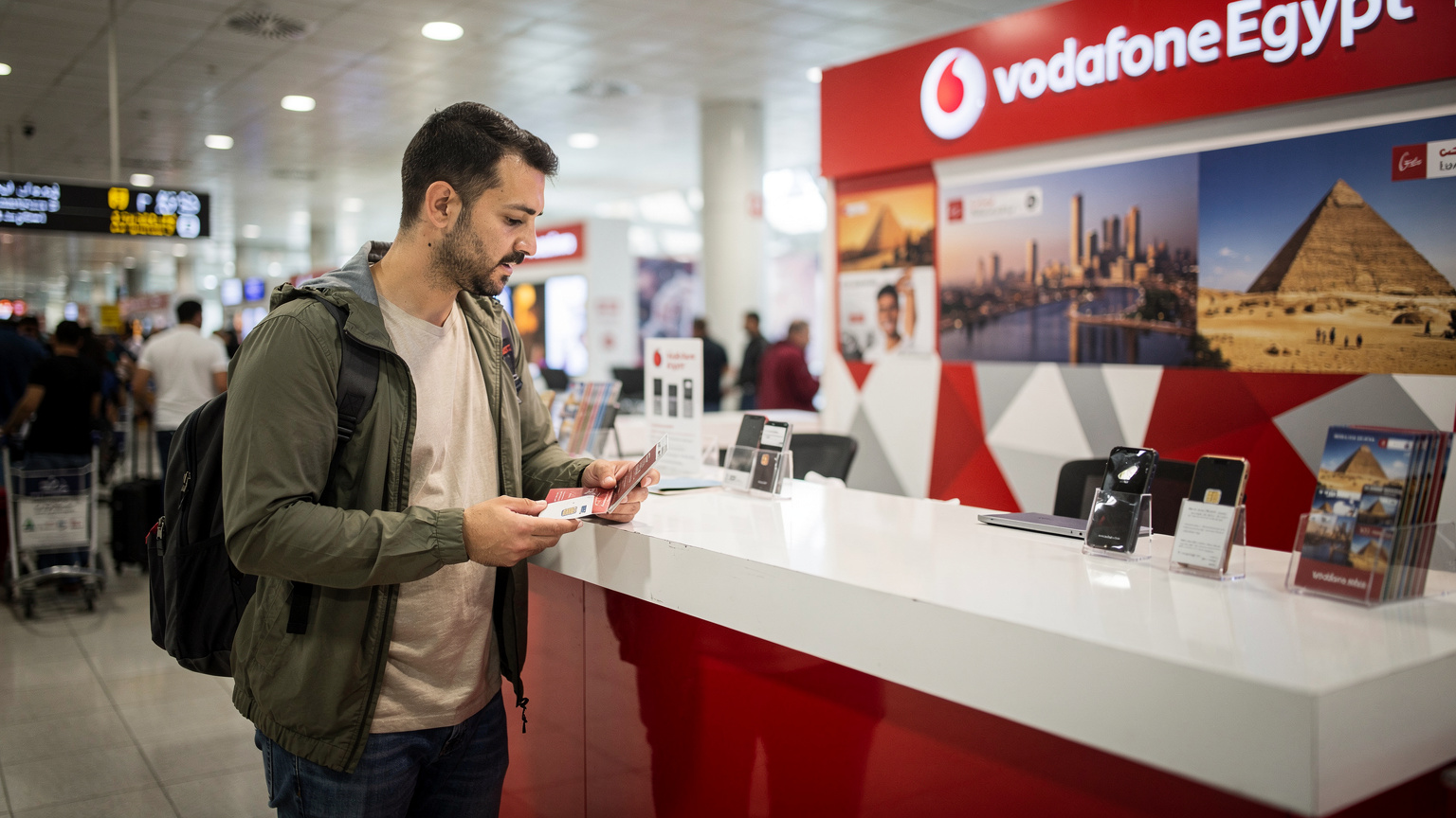 Ein Reisender wartet an einem Vodafone Egypt Stand am Flughafen Kairo, mit seinem Reisepass in der Hand, während er darauf wartet, dass seine lokale SIM-Karte aktiviert wird. Der Stand ist belebt und zeigt typische ägyptische Marketingmaterialien.
