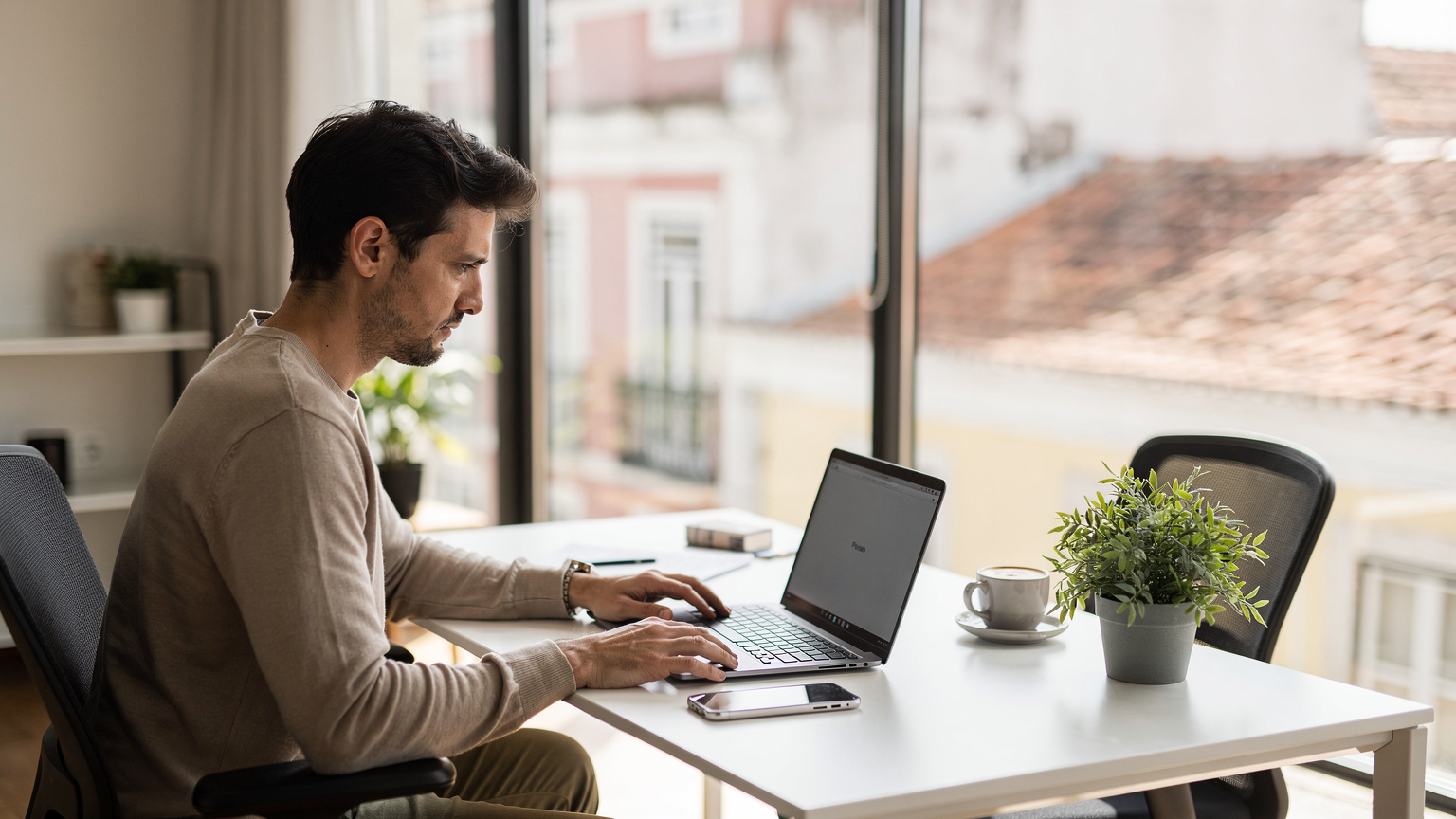 Uma pessoa sentada em uma mesa de trabalho organizada em um espaço de co-working moderno em Lisboa, com seu iPhone 18, laptop e café, trabalhando concentradamente sob a luz natural que entra pela janela.