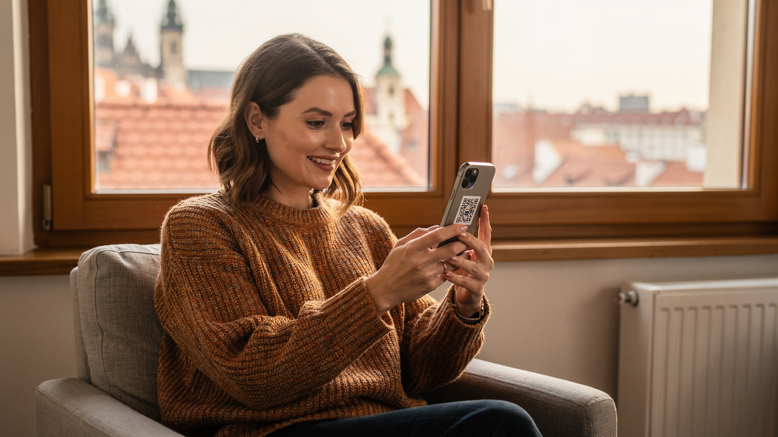 Žena s úsměvem, oblečená do podzimního svetru, naskenuje QR kód eSIM na svém iPhonu, zatímco sedí u okna s výhledem na historické střechy v Praze.