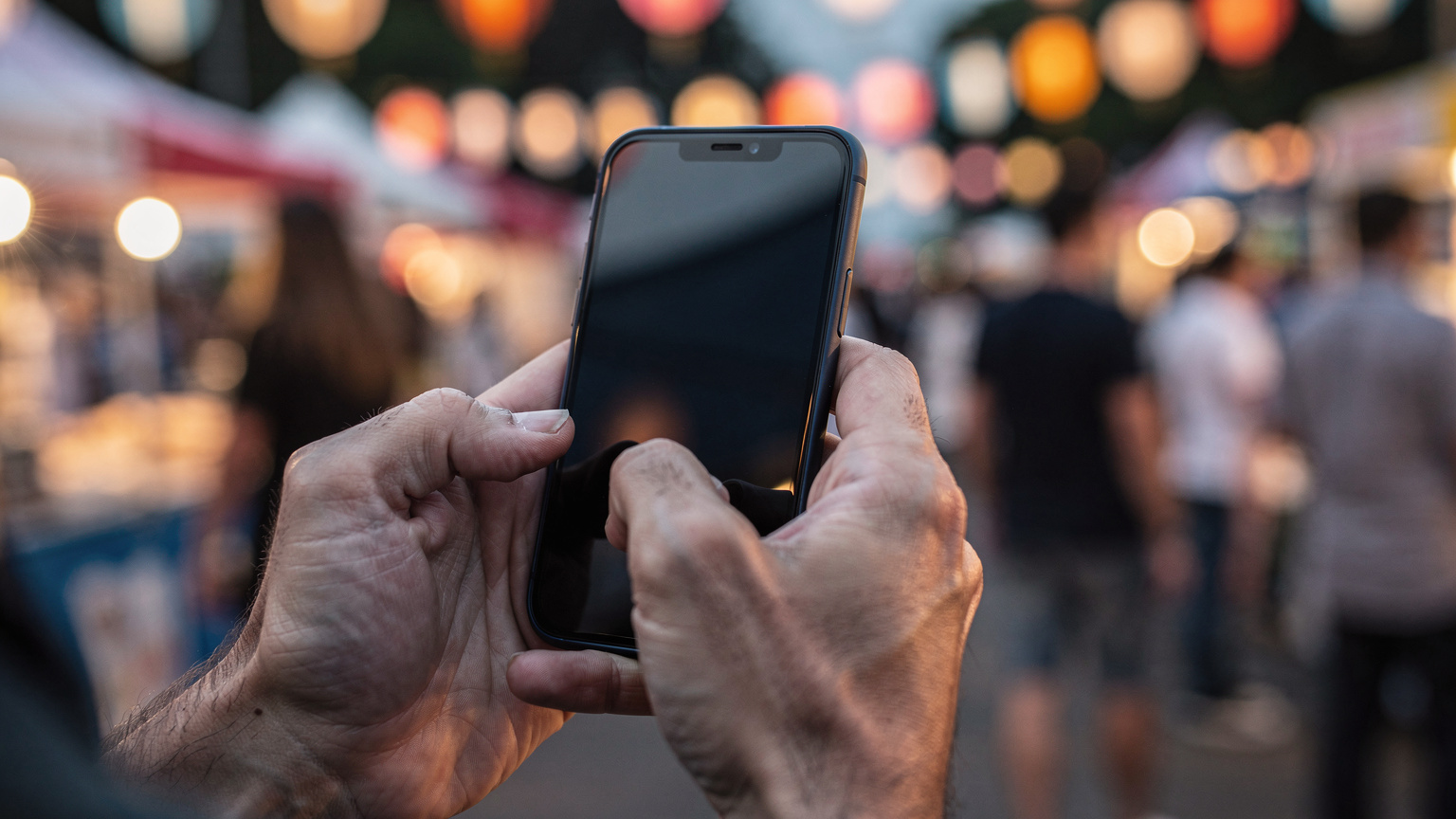 Ένας ταξιδιώτης κρατάει ένα smartphone, με το δάχτυλό του να δείχνει στην οθόνη, σε ένα περιβάλλον φεστιβάλ με θολό φόντο, ενώ προσπαθεί να βρει πληροφορίες ή να επικοινωνήσει.
