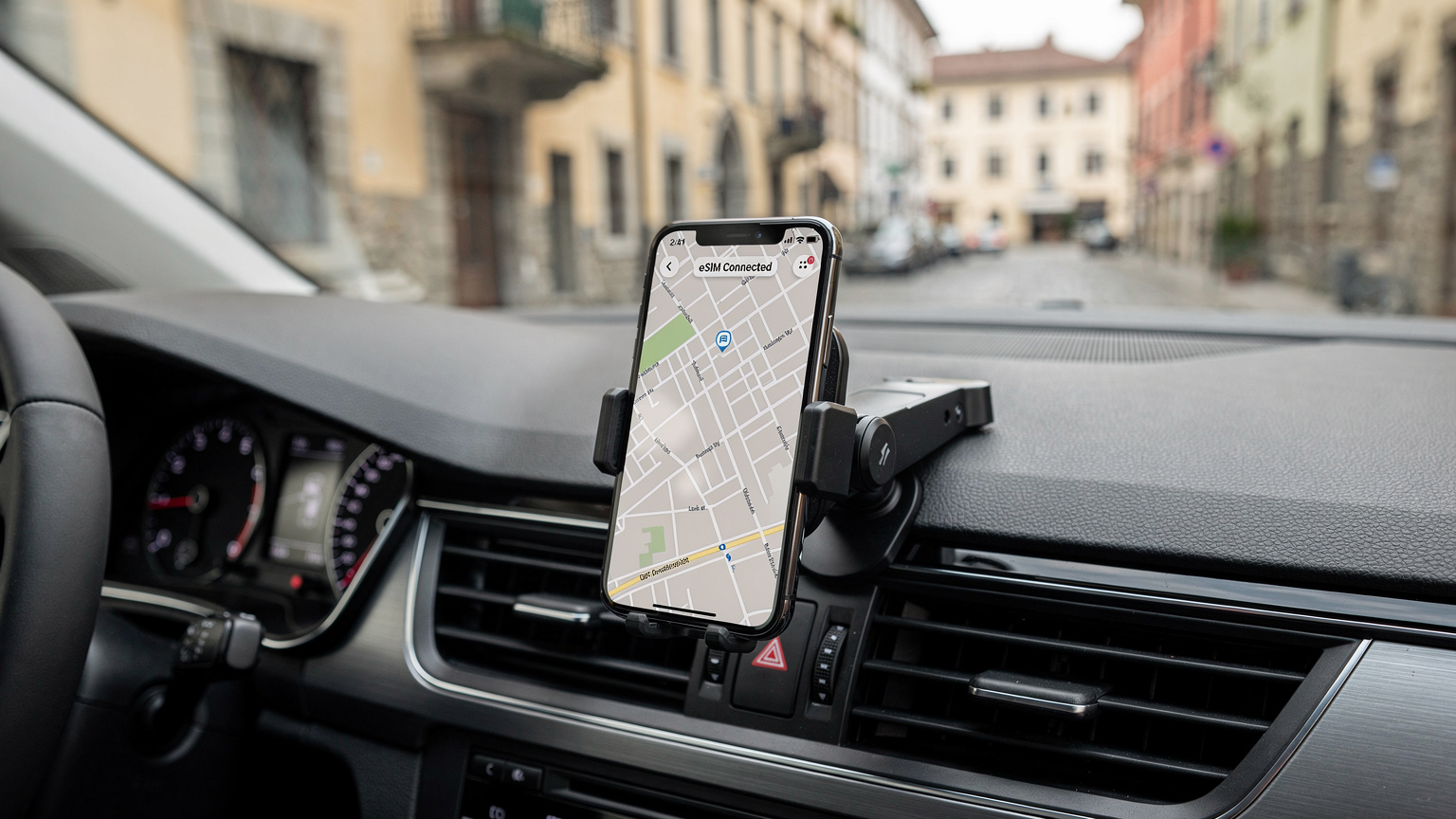 En mobiltelefon med en eSIM-profil som visar "Ansluten" i en bil, där displayen bakom visar en navigeringskarta genom en europeisk stad.