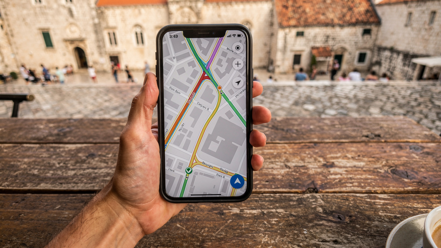 Ένα χέρι κρατά ένα smartphone με την οθόνη να δείχνει έναν χάρτη με διαδρομές, πάνω σε ένα τραπέζι καφενείου στην παλιά πόλη του Δουβροβνίκ, με φόντο την πλατεία.