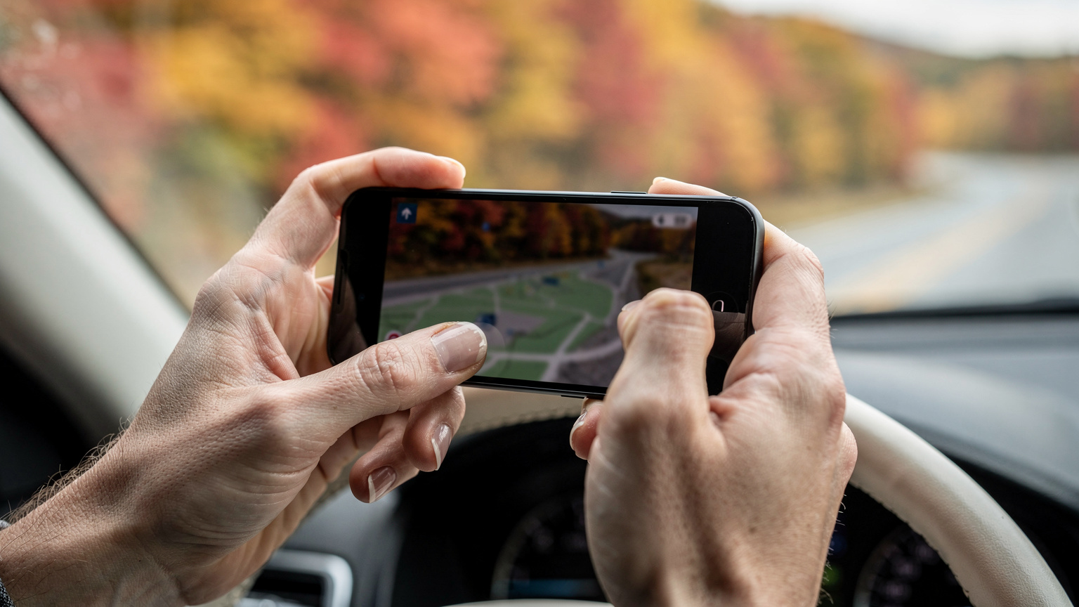 Close-up of a traveler's hands interacting with a smartphone, tapping on a digital map application to pinpoint a location while sitting in a car, with blurred autumn scenery in the background.