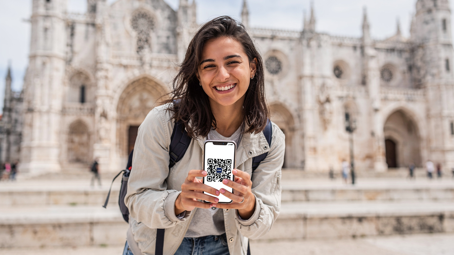 Uma estudante, recém-chegada em Portugal, sorri aliviada após ativar seu eSIM, segurando o telemóvel com o QR code de ativação na tela, em frente ao Mosteiro dos Jerónimos.