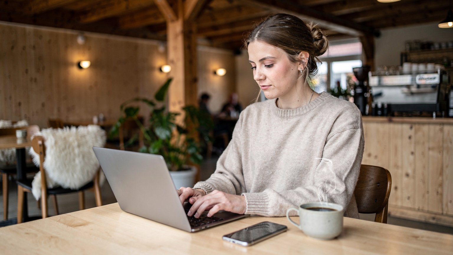 En kvinde sidder ved et minimalistisk træbord i en moderne, hyggelig norsk café, omgivet af naturligt træ og varme tekstiler. Hun arbejder fokuseret på sin laptop, med en smartphone liggende ved siden af, mens en dampende kaffekop står klar. Scenen indfanger roen ved at arbejde online i Nordens hyggelige omgivelser.