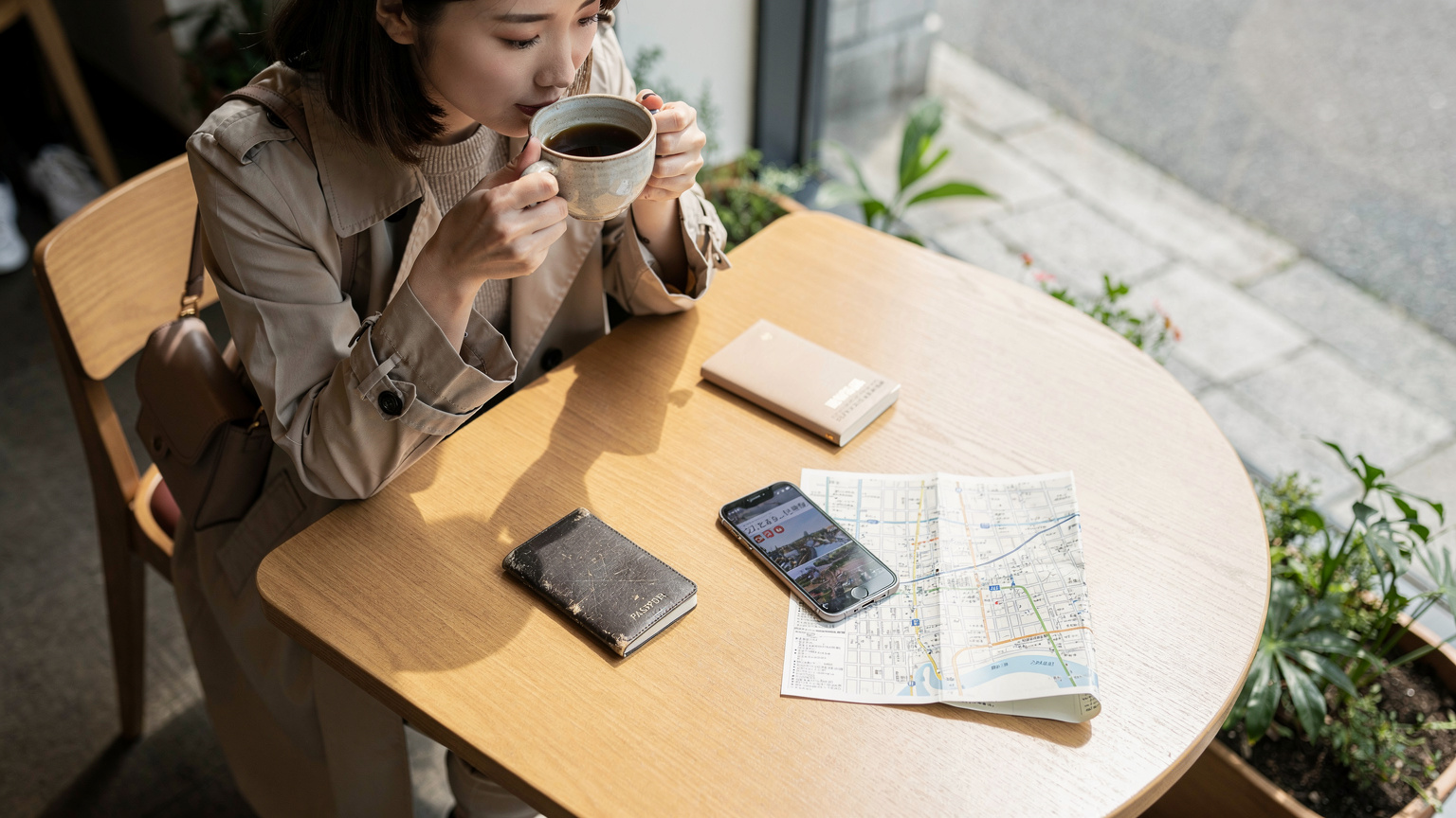 旅先のカフェで、コーヒーを飲みながらスマホで次の目的地を調べている日本人女性。手元にはパスポートやガイドブックが置かれている。