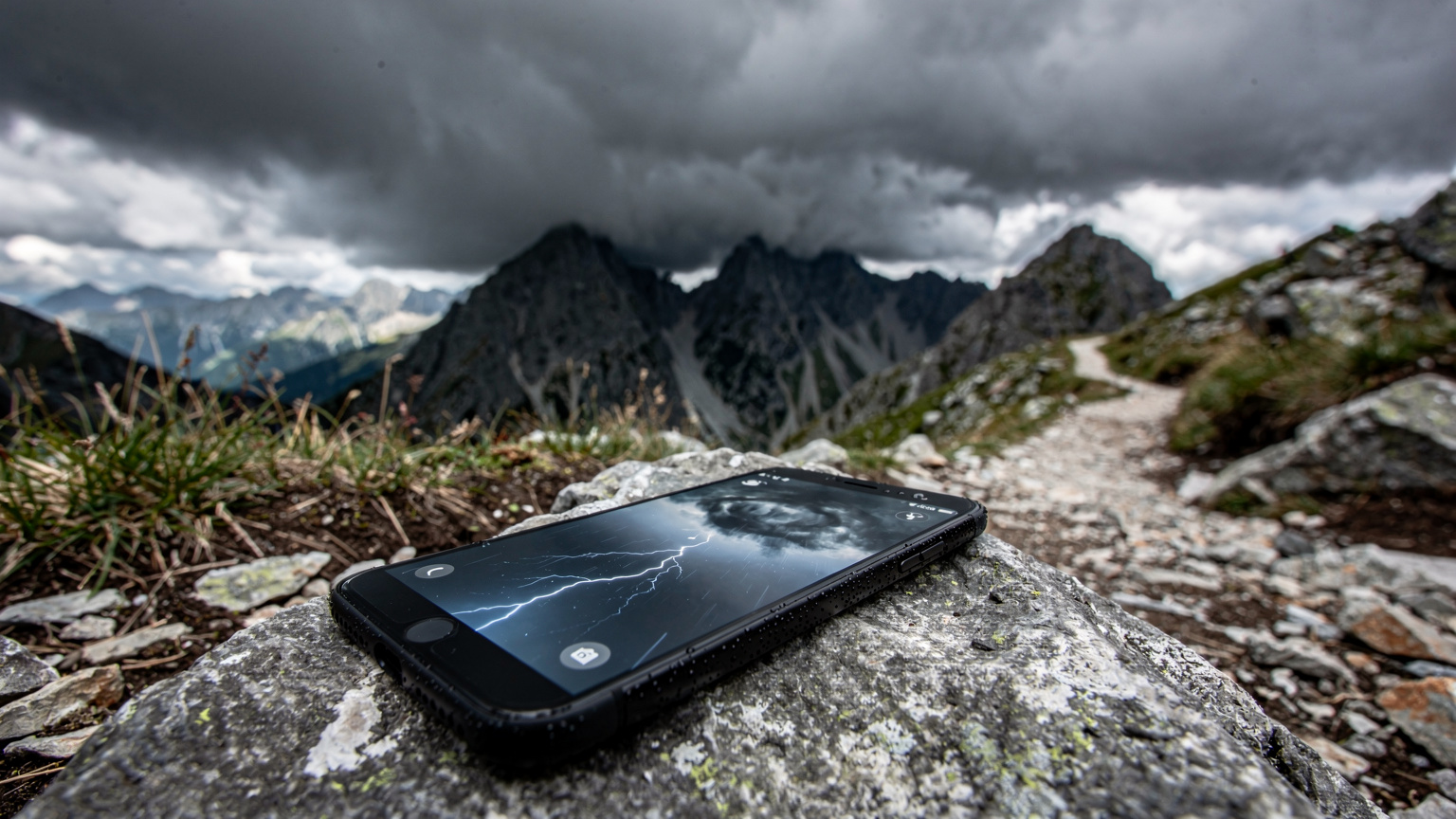 Smartfon leżący na szorstkim kamieniu na szlaku górskim, na ekranie widać aplikację pogodową z ikoną burzy, a w tle ciemne, groźne chmury zbierające się nad skalistymi szczytami Tatr.