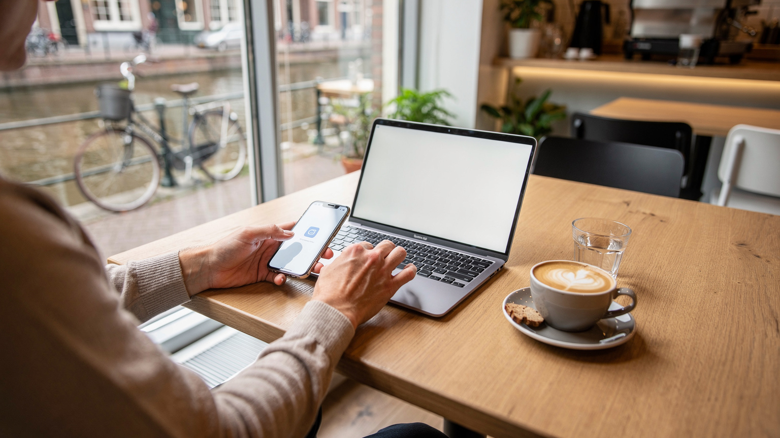 Een persoon zit aan een tafel in een modern, licht café in Amsterdam, met een kopje koffie en een laptop. Ze is geconcentreerd bezig met het instellen van een eSIM op haar smartphone, die naast de laptop ligt.