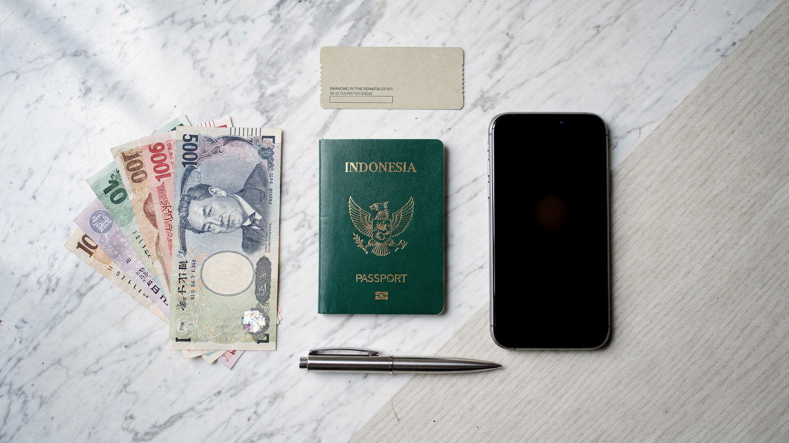 Tampilan atas paspor Indonesia, smartphone, dan beberapa mata uang asing di meja kafe yang rapi, siap untuk perjalanan.